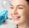In St. Augustine, FL, at Happy Smiles Orthodontics, a patient with braces smiles in the dental chair as a gloved dentist prepares to use a dental tool.