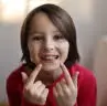 At Happy Smiles Orthodontics in St. Augustine, FL, a child in a red shirt smiles and points to their teeth for the camera indoors.
