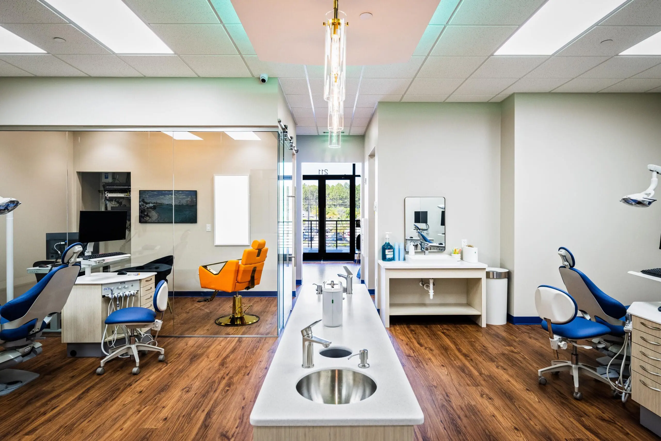 At Happy Smiles Orthodontics in St. Augustine, FL, the modern dental clinic features blue and orange exam chairs, white countertops, state-of-the-art equipment, wood-patterned flooring, glass partitions, and large windows.