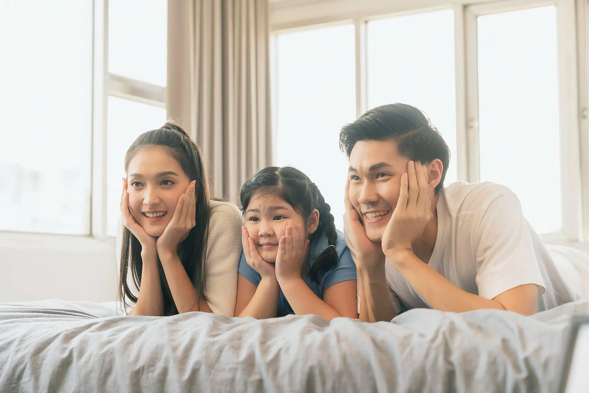 In a cheerful room with large windows, two adults and a child smile brightly while lying on a bed with their faces in their hands—showcasing the results of orthodontic appliances from Happy Smiles Orthodontics in St. Augustine, FL.