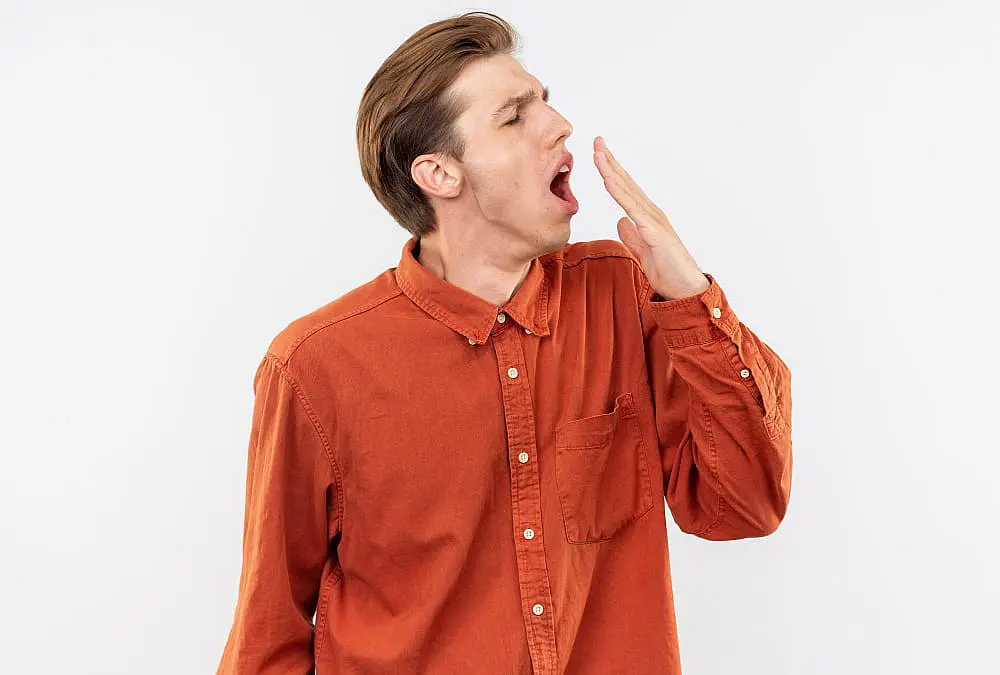 A young man, resembling a classic mouth breather, yawns with his hand over his mouth while wearing an orange button-up shirt against a plain white background—perfect for highlighting dental concerns at Happy Smiles Orthodontics in St. Augustine, FL.