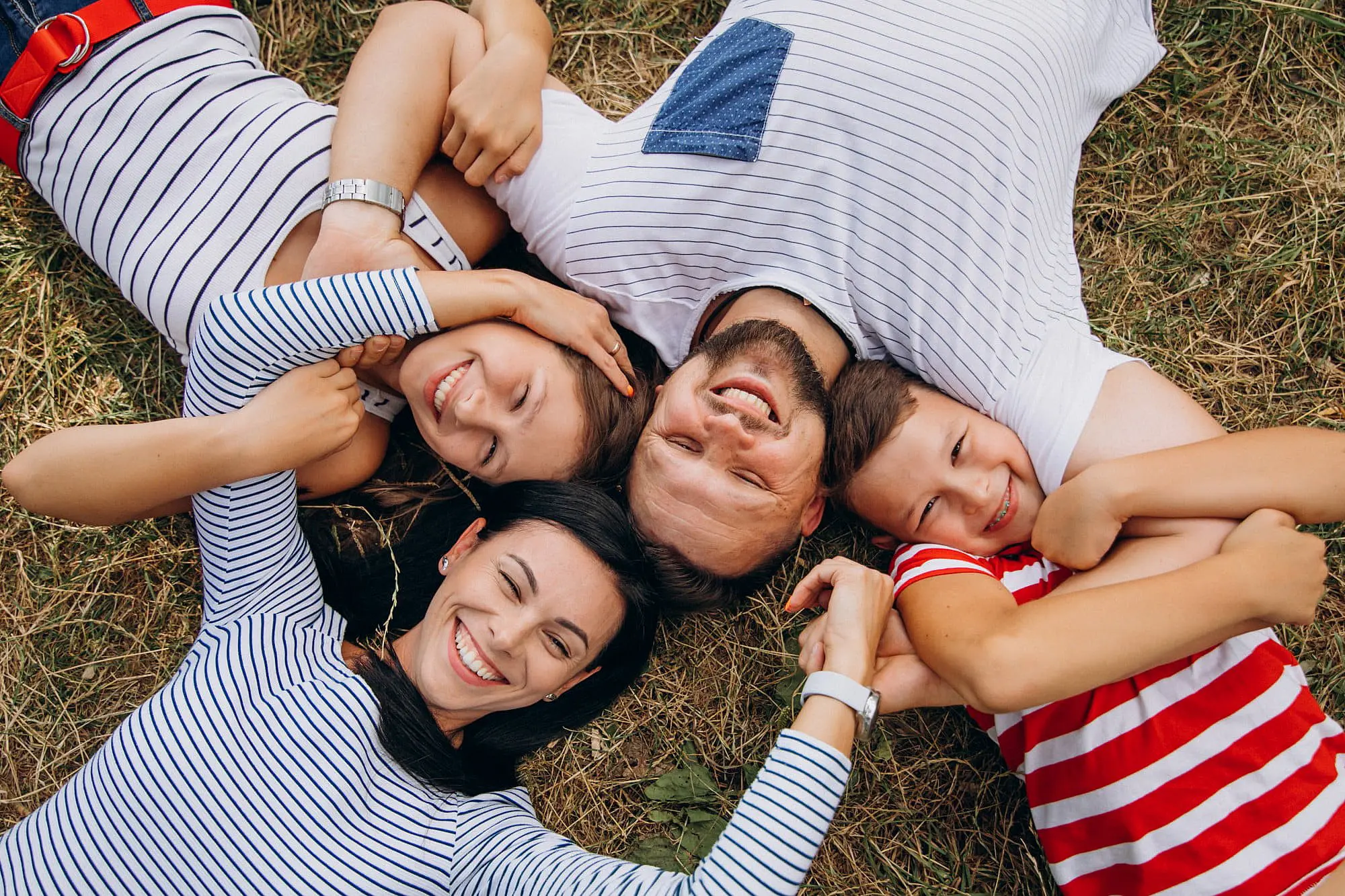 In St. Augustine, FL, a family of four in striped shirts lies on the grass in a circle with heads together, smiling for the camera—one proudly displaying braces from Happy Smiles Orthodontics.