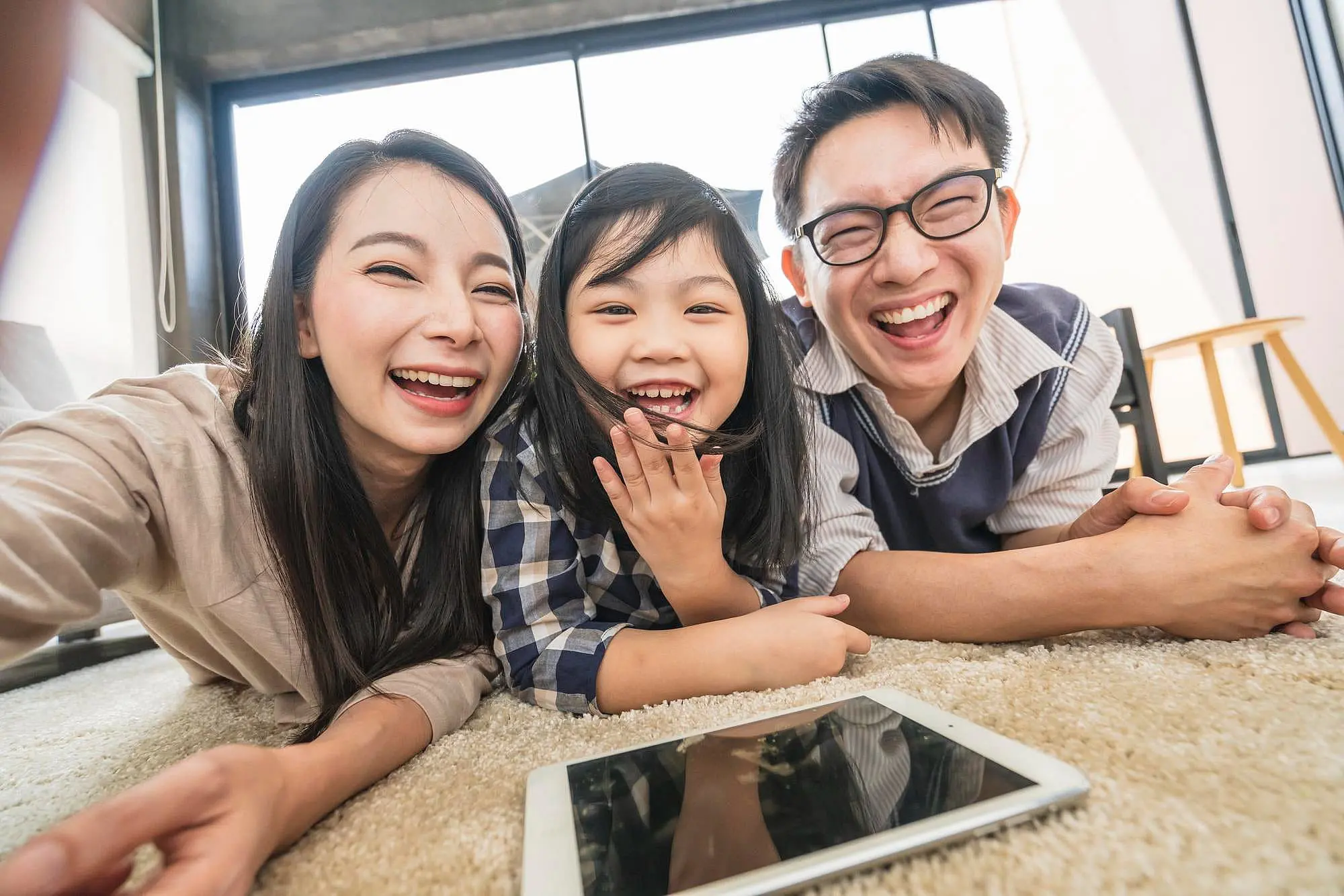 A woman, a child, and a man relax together on the floor in a bright room, smiling as they research braces cost at Happy Smiles Orthodontics in St. Augustine, FL using a tablet.