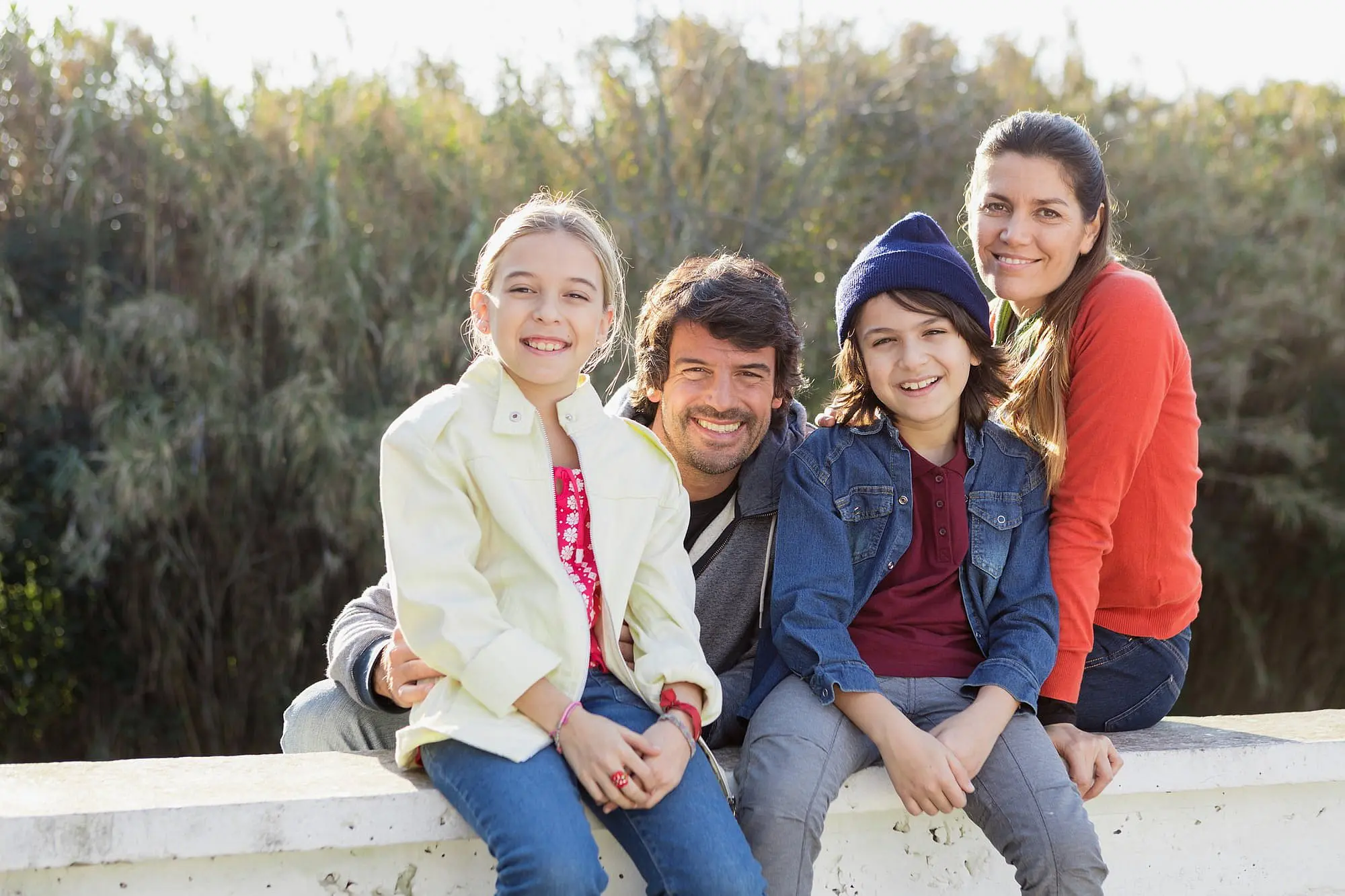 A smiling family—two adults and two children—sits together outdoors on a sunlit concrete ledge surrounded by trees, enjoying quality time while discovering different types of braces at Happy Smiles Orthodontics in St. Augustine, FL.
