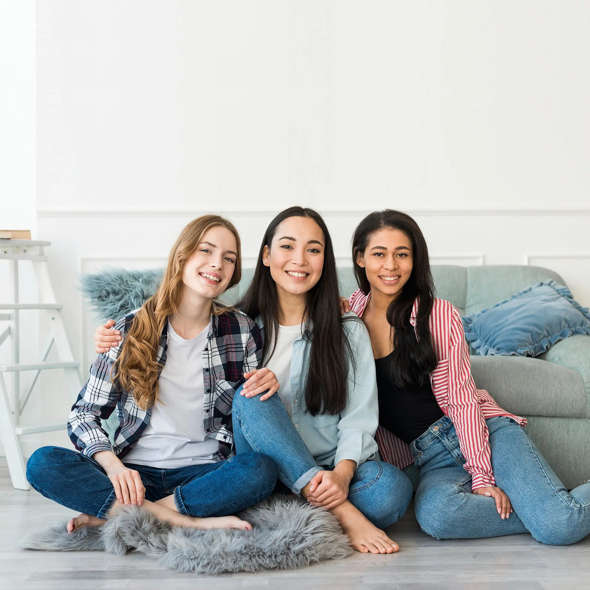 At Happy Smiles Orthodontics in St. Augustine, FL, three casually dressed young women sit together on a living room floor, smiling confidently at the camera while one proudly displays her new clear braces.