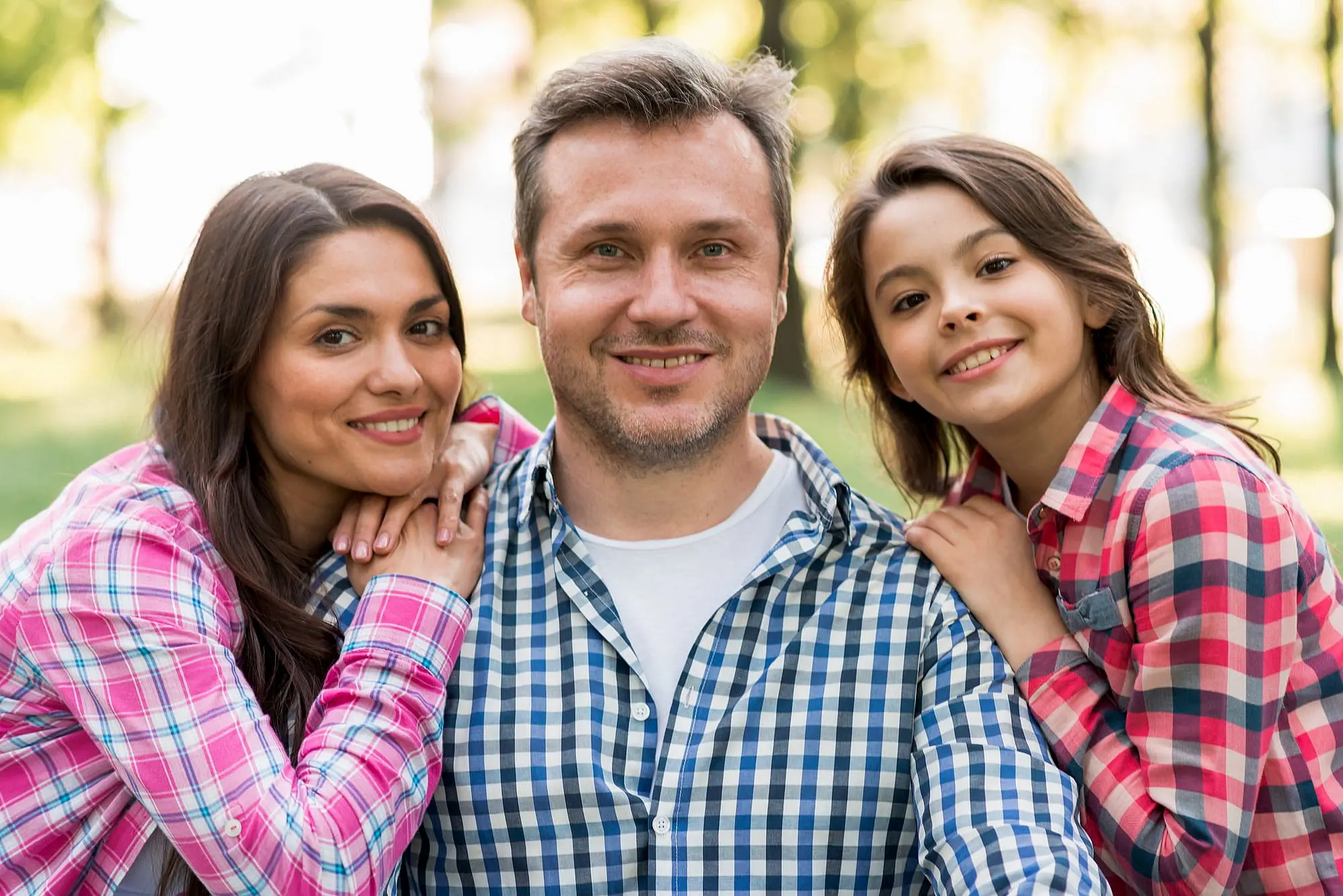A man sits outdoors in St. Augustine FL, smiling with a woman and young girl leaning close—everyone in plaid shirts and trees behind them—perhaps celebrating their new Braces Payment Plan at Happy Smiles Orthodontics.