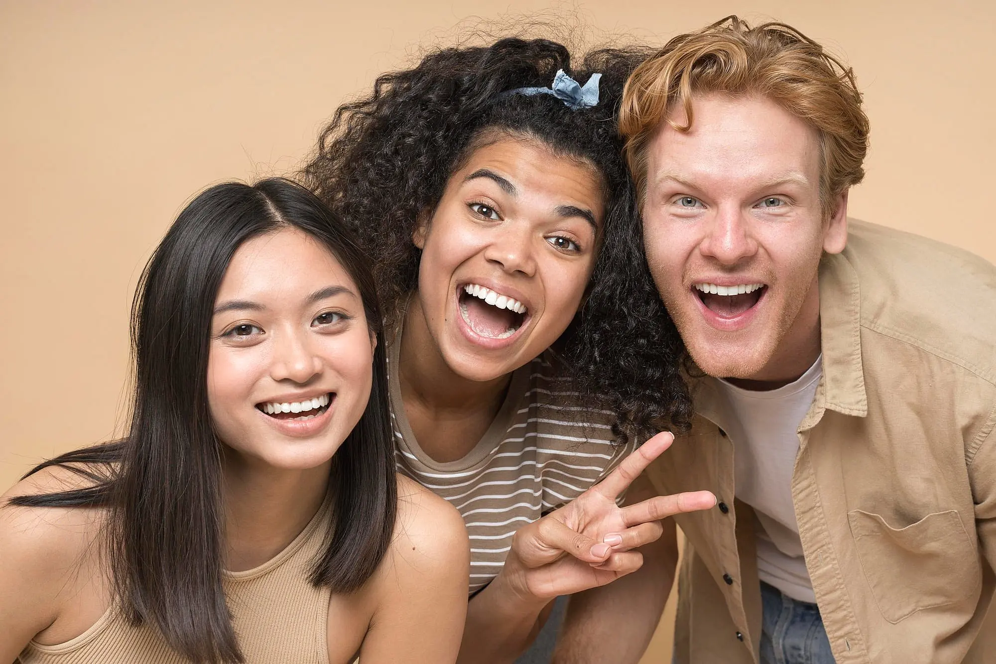 At Happy Smiles Orthodontics in St. Augustine, FL, three young adults smile for the camera as the woman in the center proudly displays her Invisalign results and flashes a peace sign—showing that confidence is possible without a high Invisalign cost.