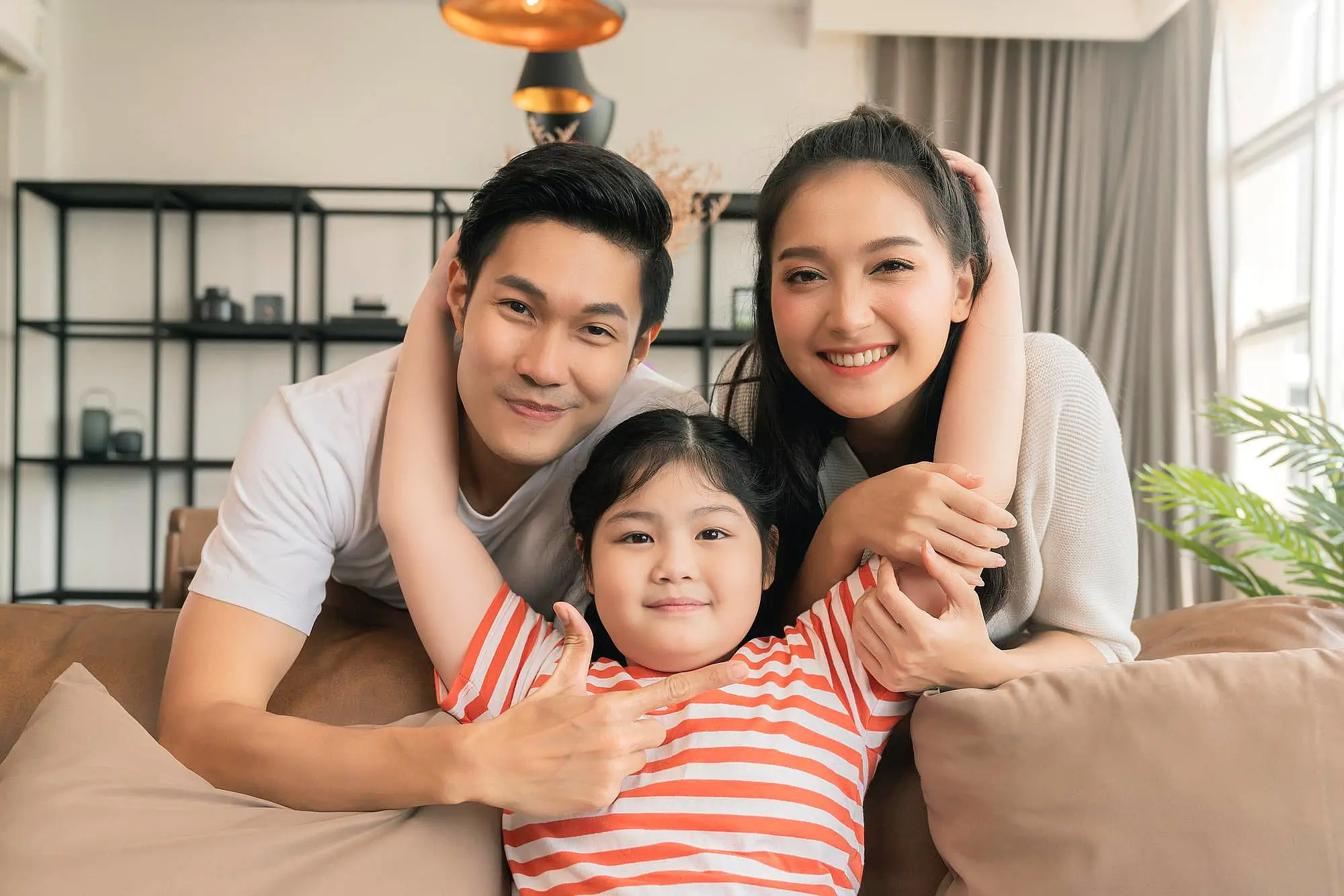 A man and woman sit on a couch with a smiling young girl in a red striped shirt, all facing the camera in a bright living room—capturing the kind of perfect smiles patients achieve at Happy Smiles Orthodontics in St. Augustine, FL when choosing Invisalign or braces.