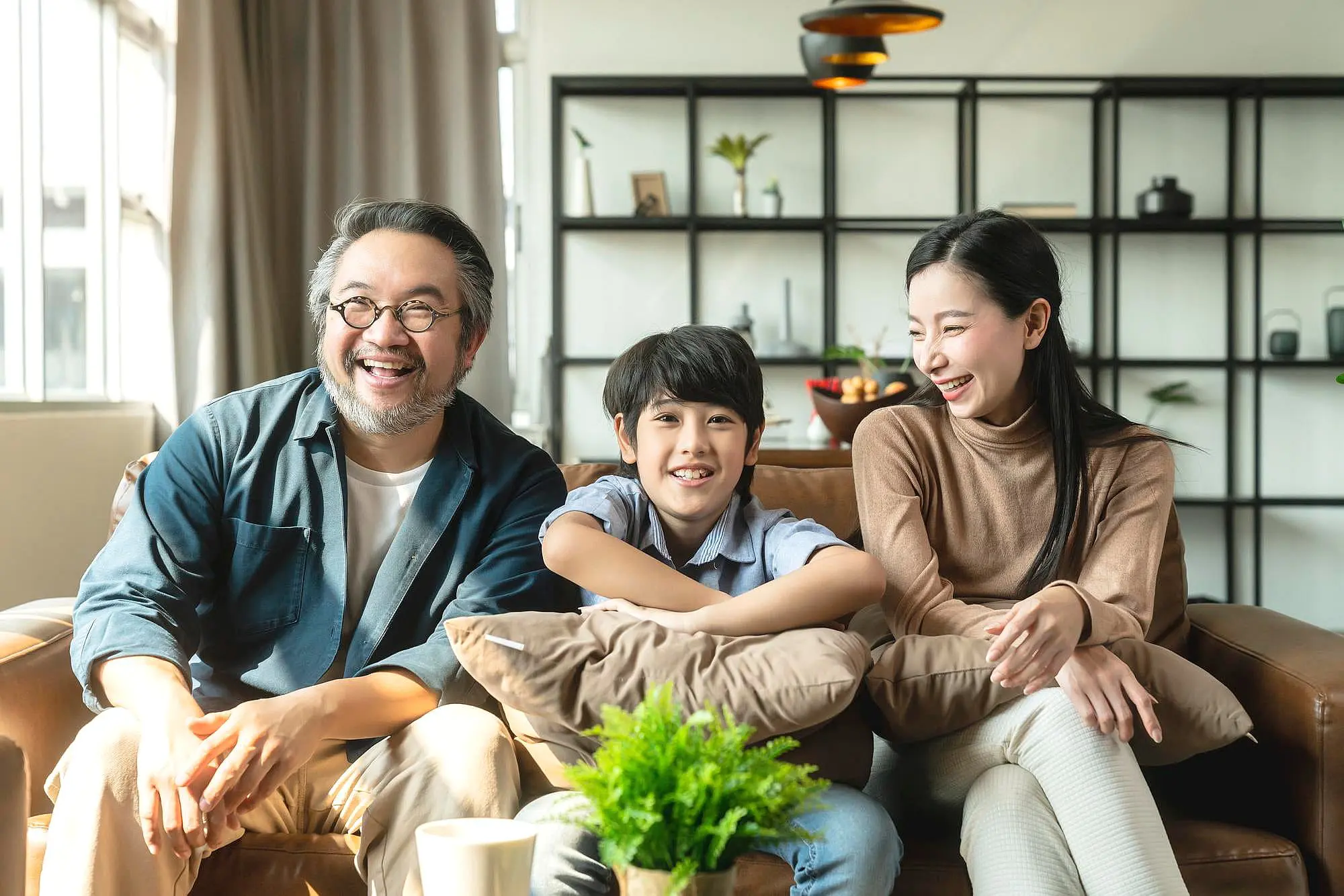 At Happy Smiles Orthodontics in St. Augustine FL, three people relax on a couch, smiling and laughing—possibly after talking about Teeth Retainer Replacement. A plant decorates the table while shelves with décor complete the living room background.