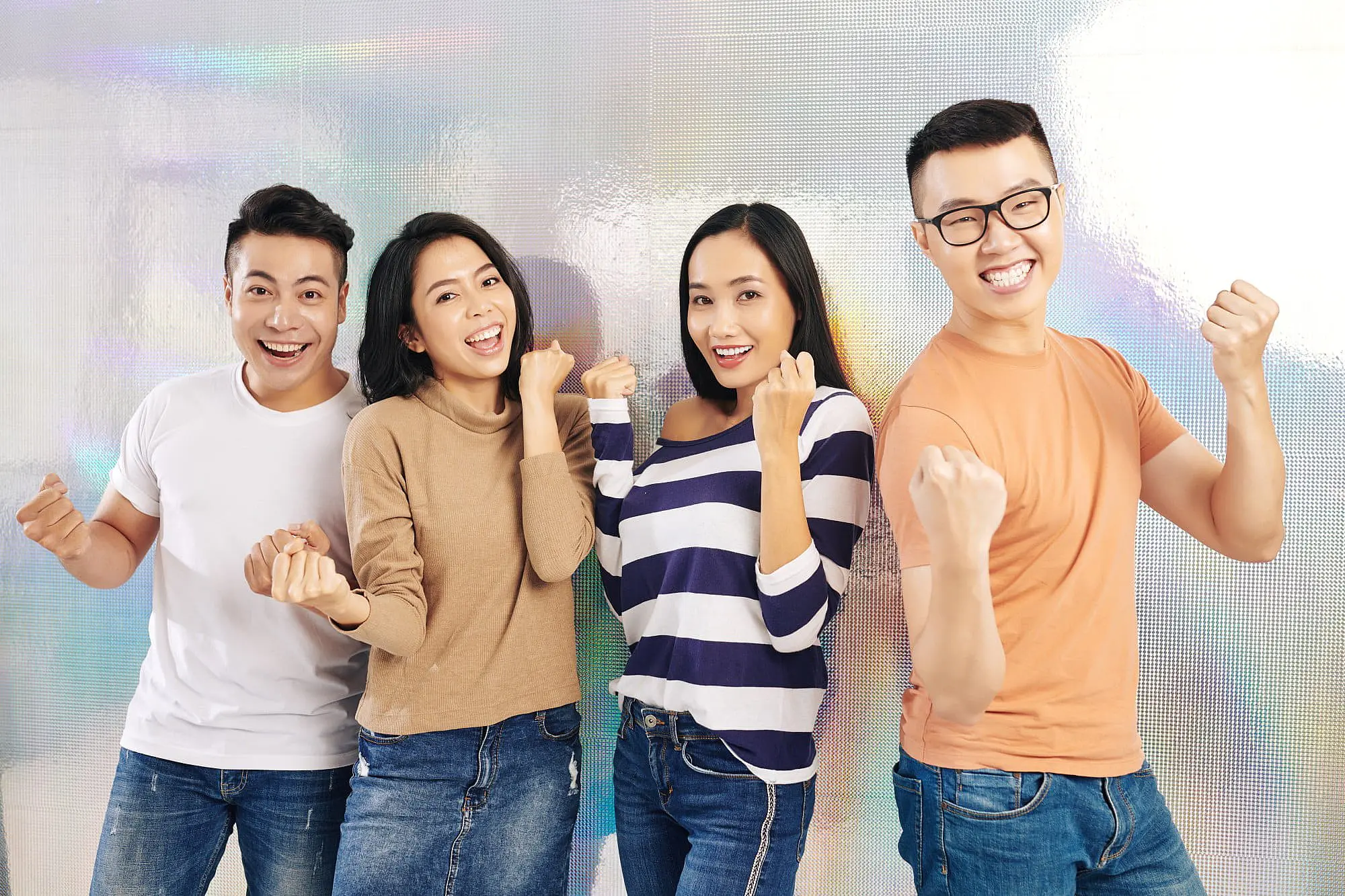Four young adults stand side by side, smiling and raising their fists triumphantly against a light textured background, perfectly capturing the spirit of Happy Smiles Orthodontics for anyone searching for “Invisalign Near Me” in St. Augustine, FL.