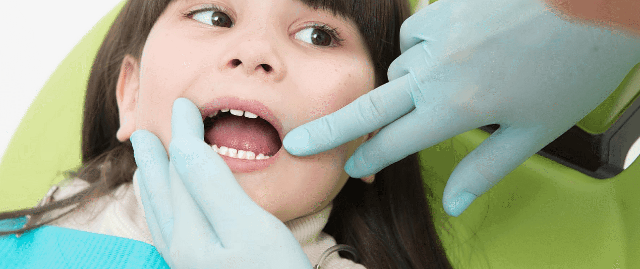 At Happy Smiles Orthodontics in St. Augustine, FL, a children’s orthodontist wearing blue gloves examines a young child’s teeth as the child sits in the dental chair with their mouth open.
