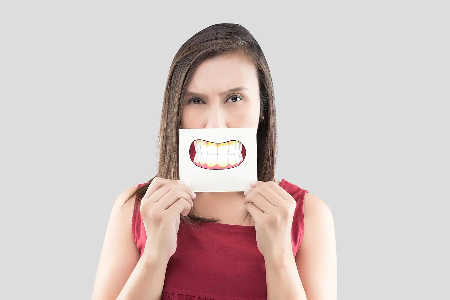 At Happy Smiles Orthodontics in St. Augustine, FL, a woman in a red sleeveless top holds a paper with a cartoon underbite smile over her mouth against a plain gray background.
