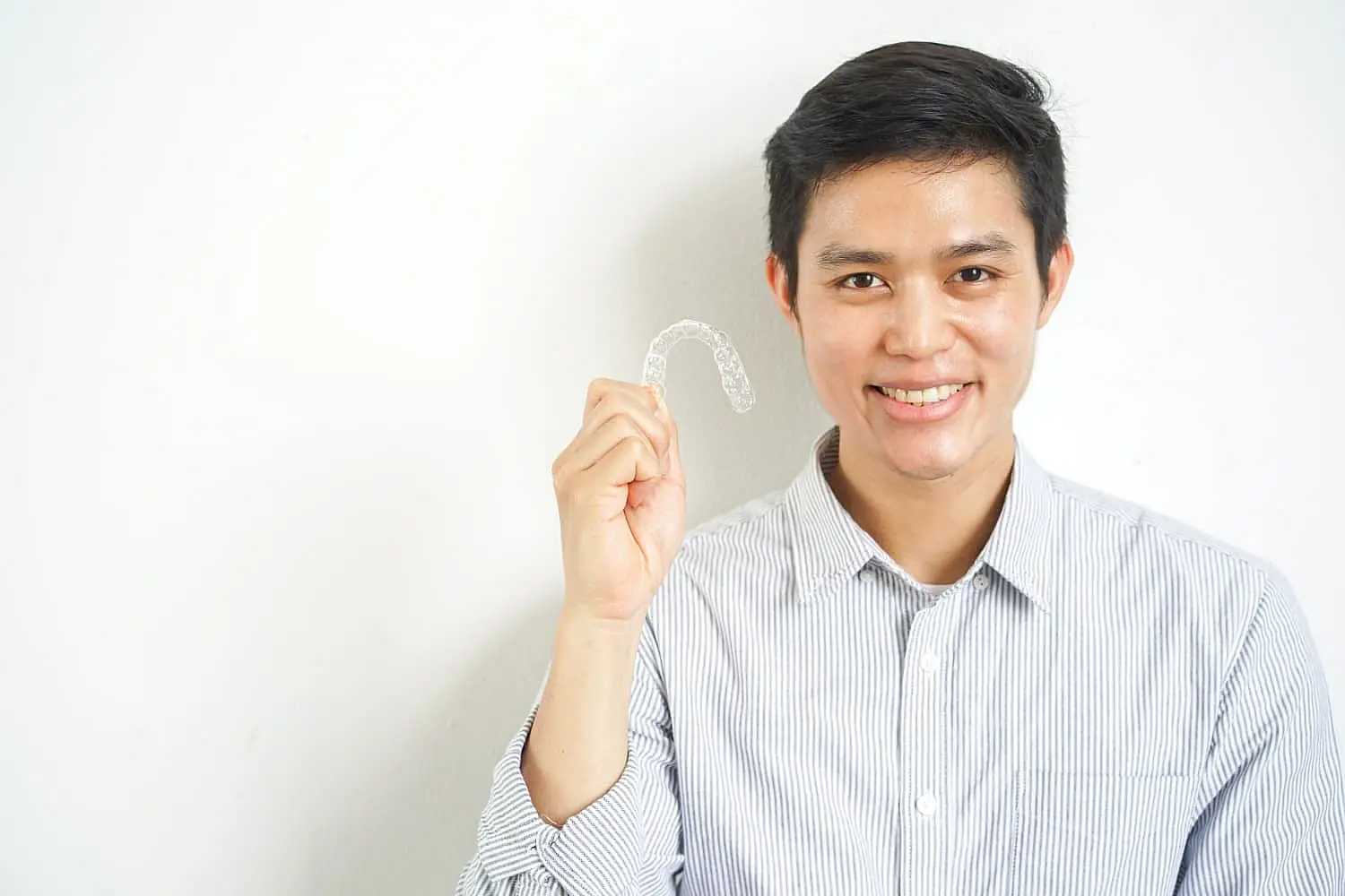 Smiling and holding a clear aligner, a person with short dark hair in a striped shirt showcases the advantages of an Invisalign Payment Plan at Happy Smiles Orthodontics in St. Augustine, FL, set against a plain white background.