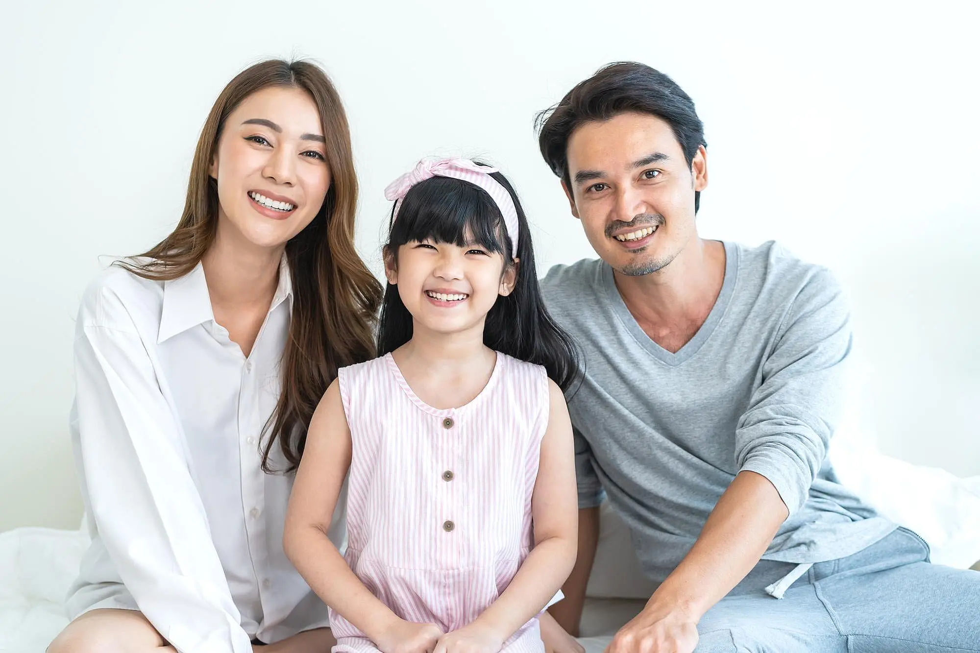 A woman, a young girl, and a man sit indoors smiling at the camera against a plain white background—resembling families who visit Happy Smiles Orthodontics in St. Augustine, FL to explore Invisalign payment plans and achieve confident smiles together.