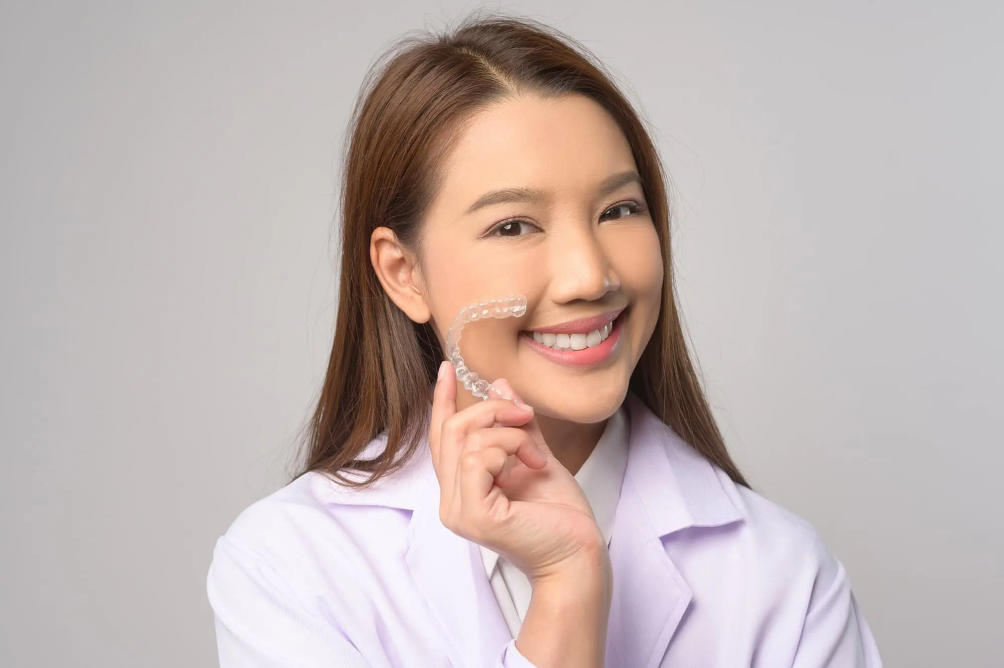 Showcasing modern adult orthodontics at Happy Smiles Orthodontics in St. Augustine, FL, a woman in a white lab coat smiles while holding an Invisalign® Clear Aligner against a plain background.