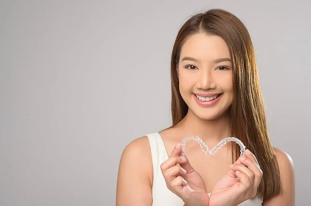 A young woman smiles at the camera, holding clear aligners shaped into a heart to highlight the benefits of an Invisalign Payment Plan with Happy Smiles Orthodontics in St. Augustine, FL.