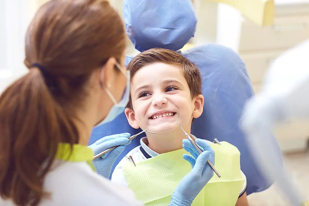 At Happy Smiles Orthodontics in St. Augustine, FL, a children’s orthodontist wearing protective masks and gloves examines a young boy's teeth as he sits comfortably in the dental chair, smiling.
