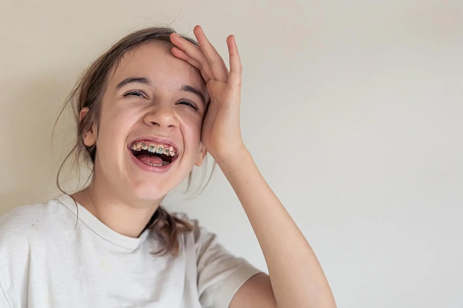 A young girl with affordable braces smiles and laughs, hand to her forehead, against a plain light background at Happy Smiles Orthodontics in St. Augustine, FL.
