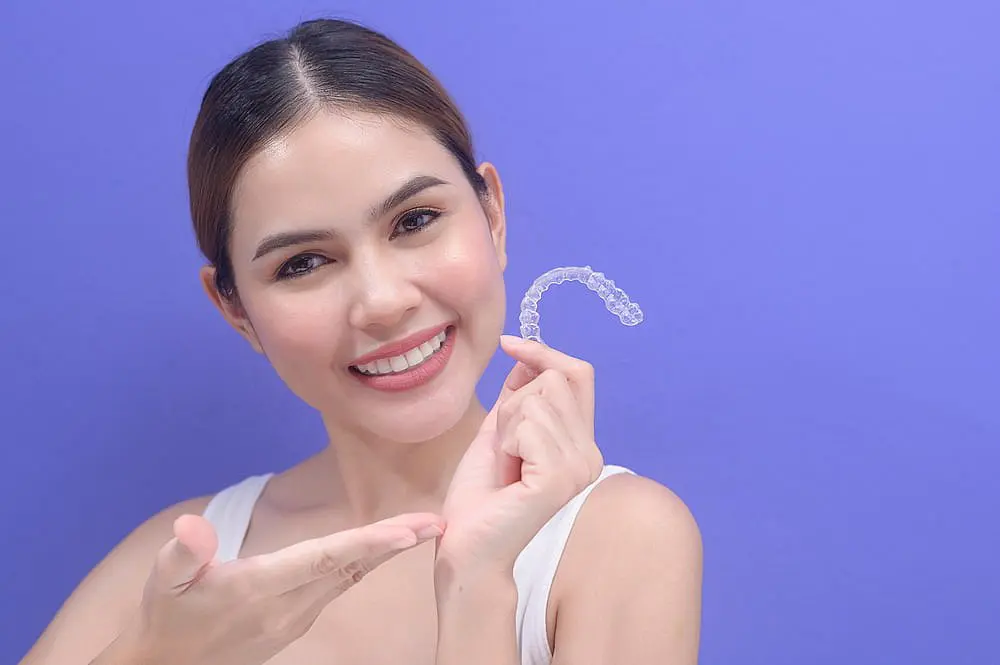 Against a purple background, a woman smiles and points to a clear aligner, showcasing how Invisalign at Happy Smiles Orthodontics in St. Augustine, FL may be covered by insurance.