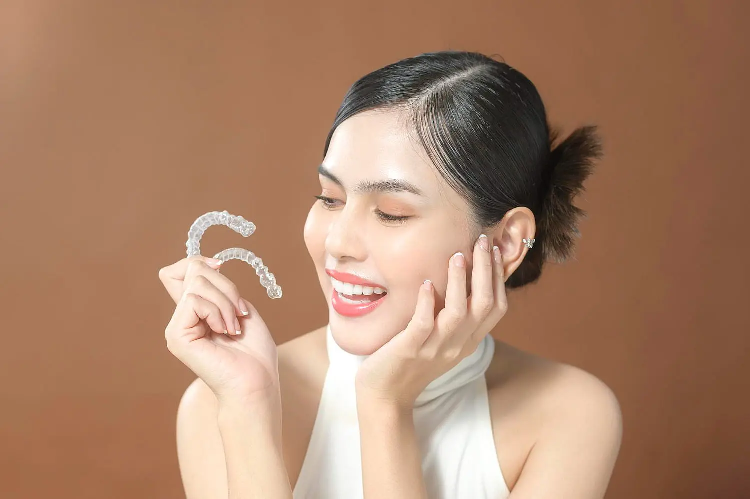 A woman smiles and admires clear dental aligners in her hand, highlighting the advantages of an Invisalign Payment Plan at Happy Smiles Orthodontics in St. Augustine, FL, set against a simple brown background.