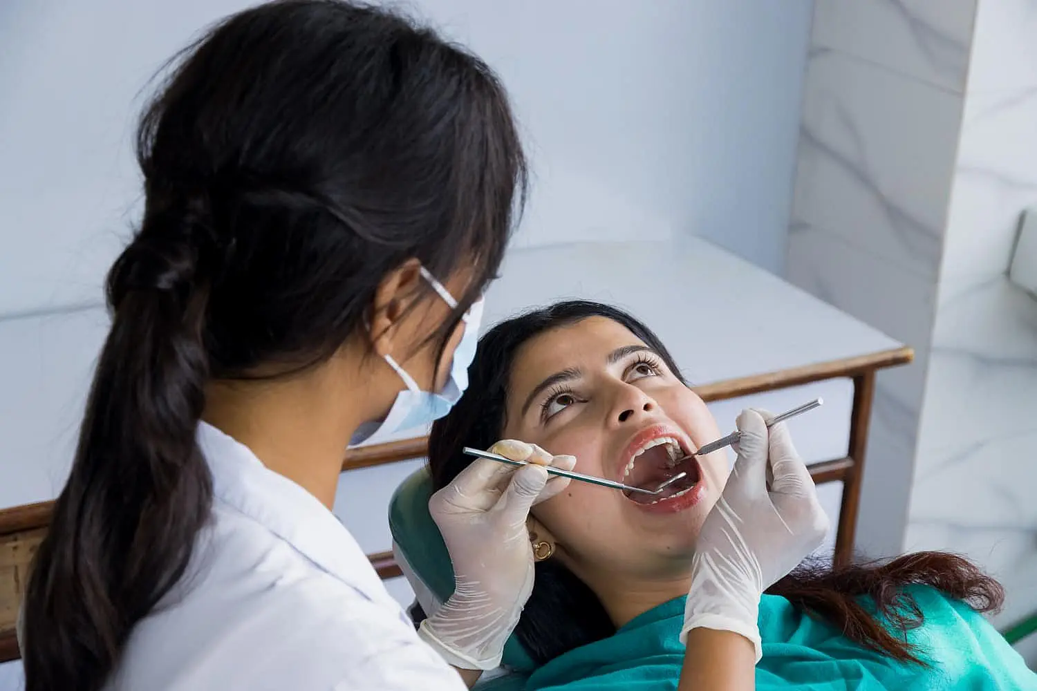At Happy Smiles Orthodontics in St. Augustine, FL, a dentist wearing gloves and a mask uses dental tools to examine a female patient’s mouth for issues like tongue thrust.