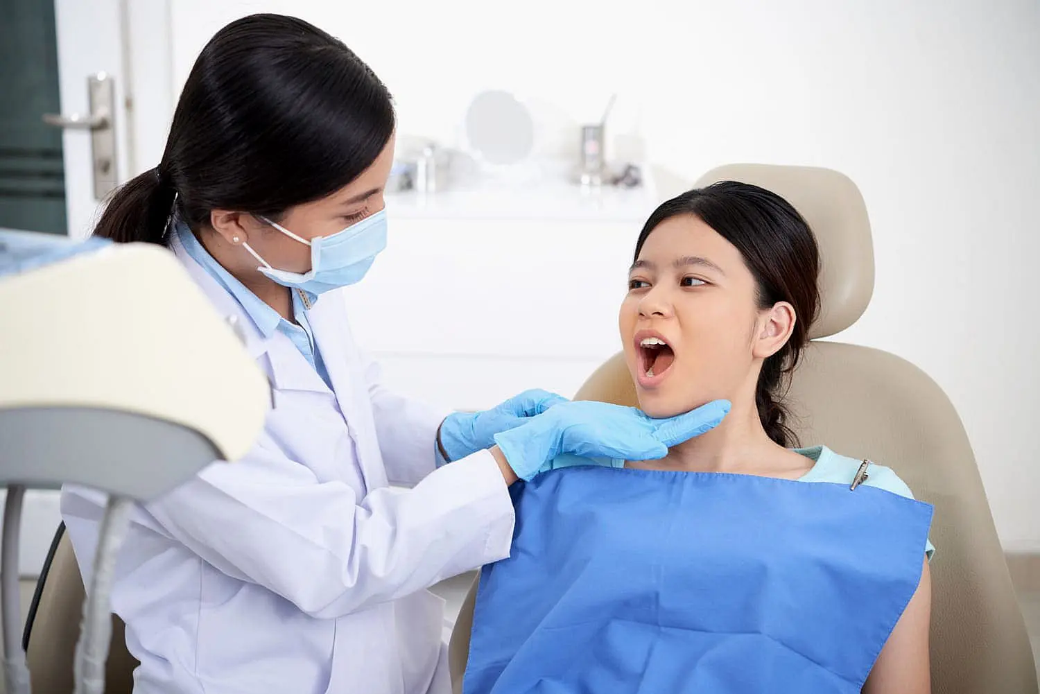 At Happy Smiles Orthodontics in St. Augustine, FL, a dentist wearing gloves and a mask examines the open mouth of a young mouth breather, who is seated and covered with a blue bib in the clinic.