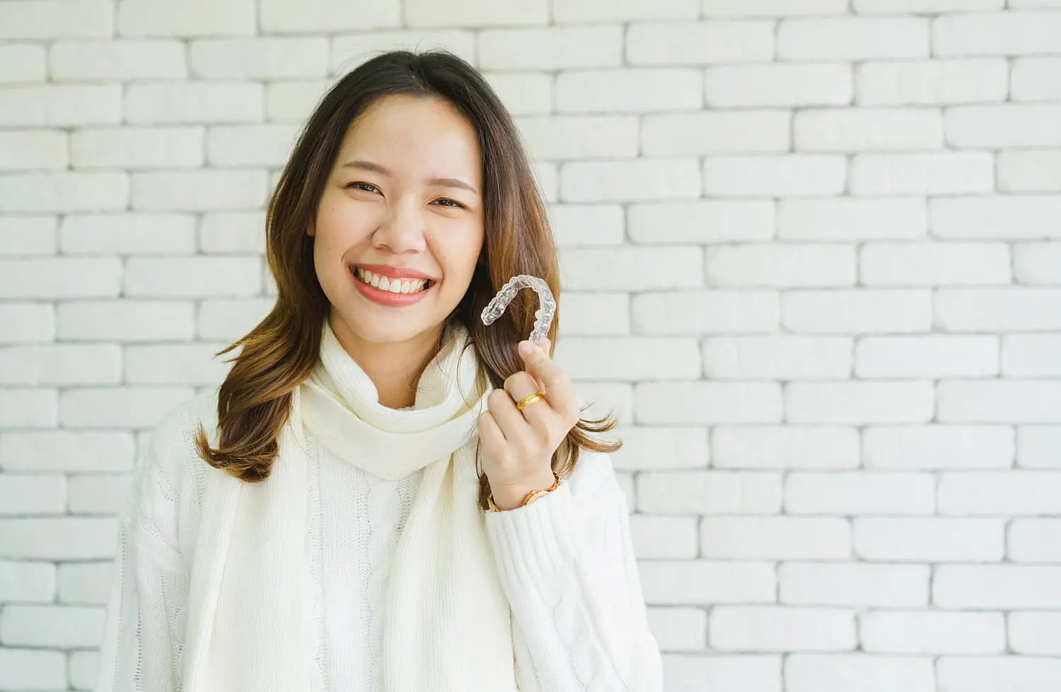 A woman in a white sweater smiles while holding a clear dental aligner, contemplating whether Invisalign is covered by insurance at Happy Smiles Orthodontics in St. Augustine, FL, with a white brick wall behind her.
