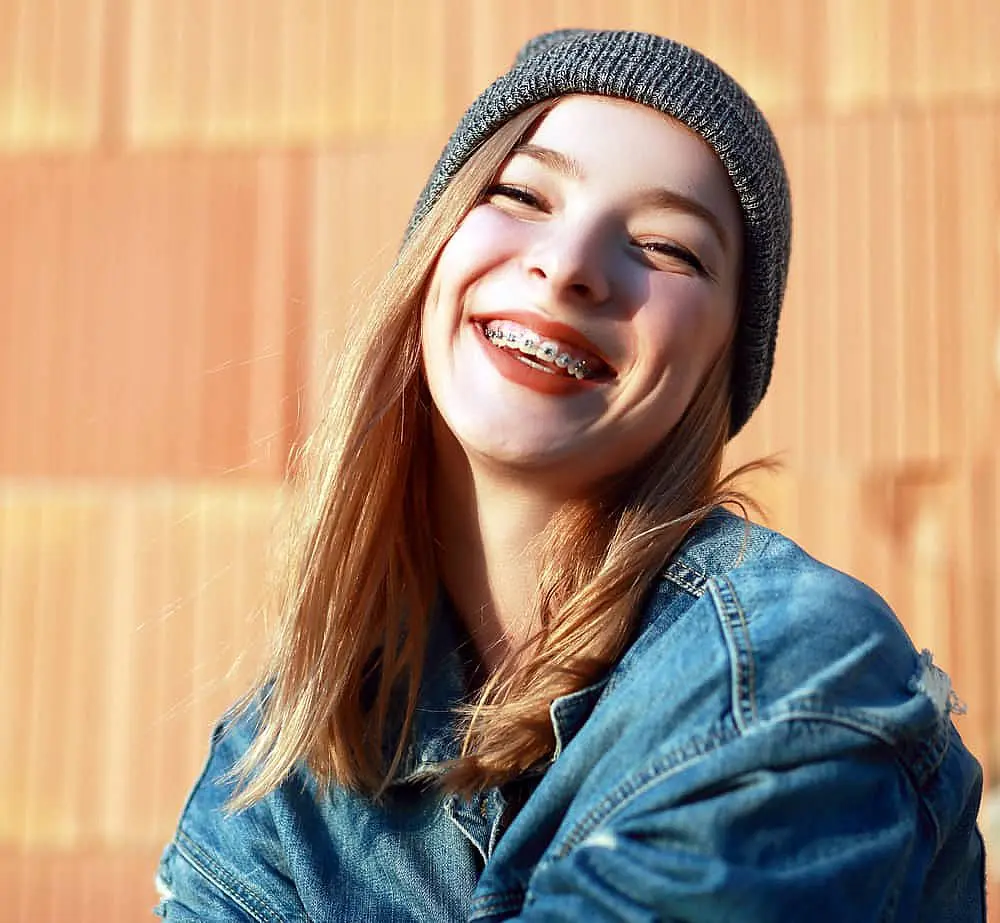 A smiling young person with straight blonde hair, a gray beanie, and a denim jacket displays metal braces in front of a textured orange wall—representing Happy Smiles Orthodontics in St. Augustine, FL.