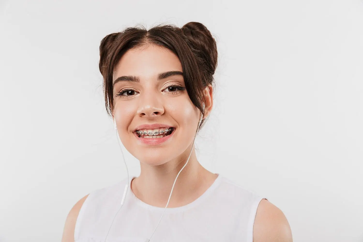 A teen girl with brown hair in two buns, wearing a white sleeveless top, metal braces, and earphones, smiles against a plain white background for Happy Smiles Orthodontics in St. Augustine, FL.