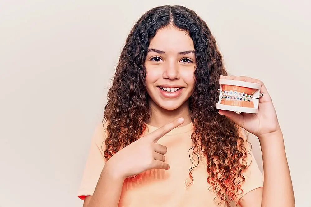Happy Smiles Orthodontics At Happy Smiles Orthodontics in St. Augustine, FL, a curly-haired girl smiles and points to a dental model with braces, showcasing orthodontic treatment and emphasizing the importance of understanding braces cost.
