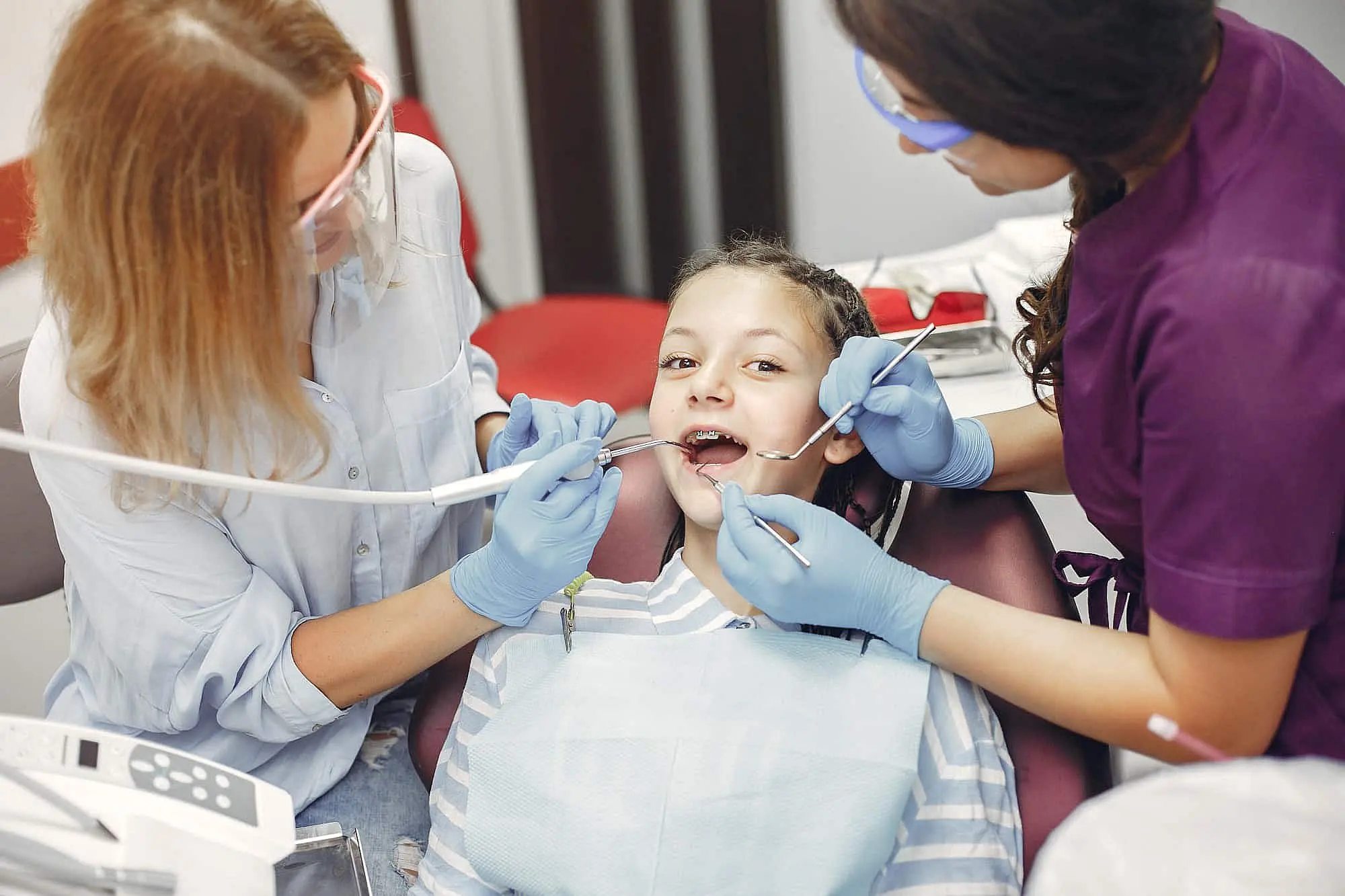 At Happy Smiles Orthodontics in St. Augustine, FL, two children's orthodontists wearing gloves and protective glasses examine a child's teeth as she sits in the dental chair with her mouth open.