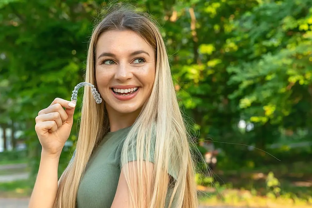 Showcasing the confidence that orthodontic insurance can offer, a woman with long blonde hair smiles while holding a clear aligner outdoors at Happy Smiles Orthodontics in St. Augustine, FL.