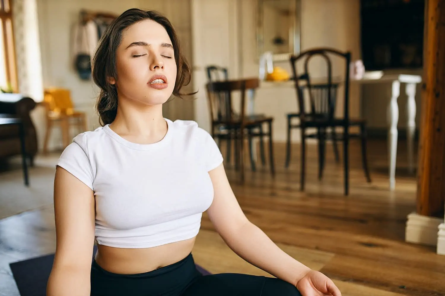 At Happy Smiles Orthodontics in St. Augustine, FL, a woman in a white crop top sits cross-legged on a yoga mat indoors, eyes closed in meditation—an ideal pose for mouth breathers seeking relaxation.