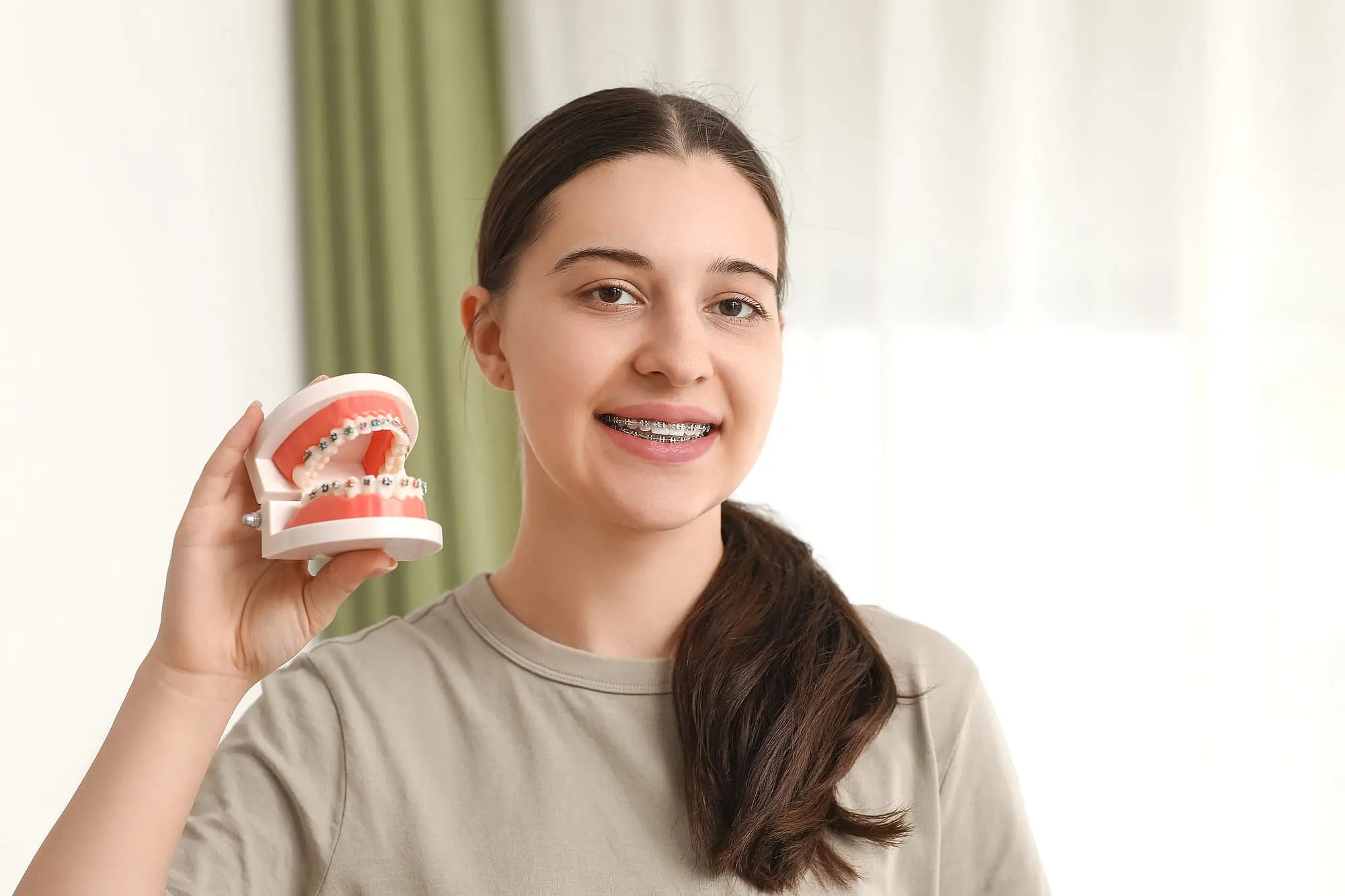 At Happy Smiles Orthodontics in St. Augustine, FL, a young woman with dark hair and braces who has an underbite holds a dental model with braces, standing indoors before a light background and green curtain.