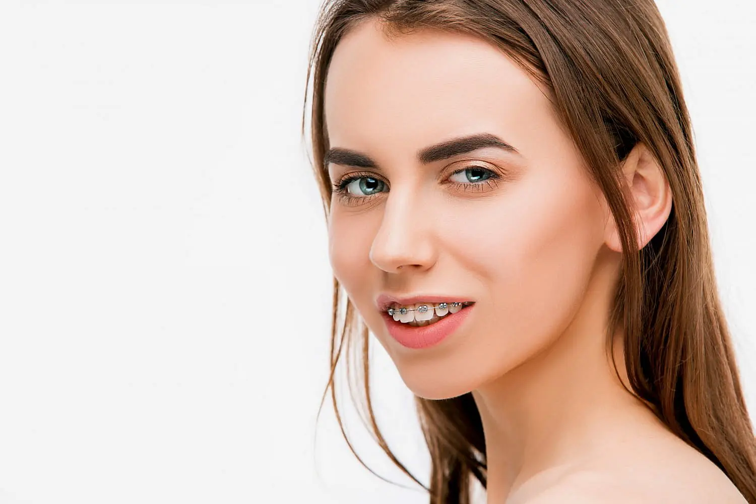 At Happy Smiles Orthodontics in St. Augustine, FL, a young woman with long brown hair and light skin smiles slightly, displaying metal braces that are correcting her overbite against a plain white background.