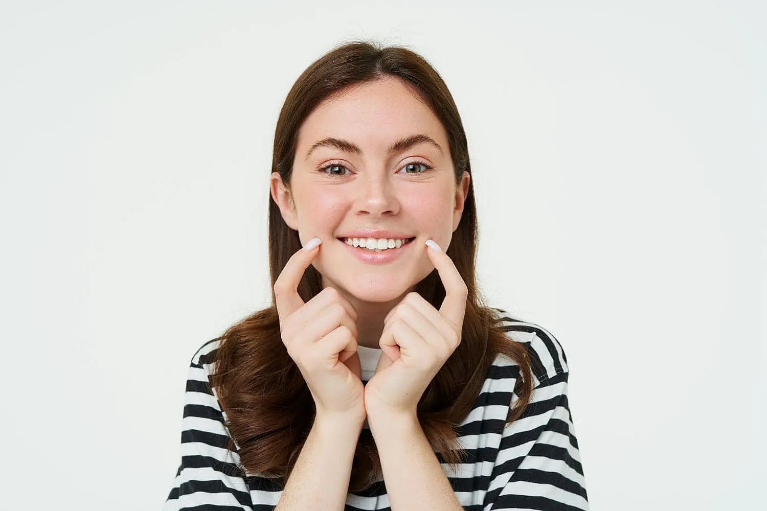 A young woman with long brown hair, wearing a striped shirt, confidently points to her teeth and smiles—showing off her smile despite a slight overbite—at Happy Smiles Orthodontics in St. Augustine, FL.