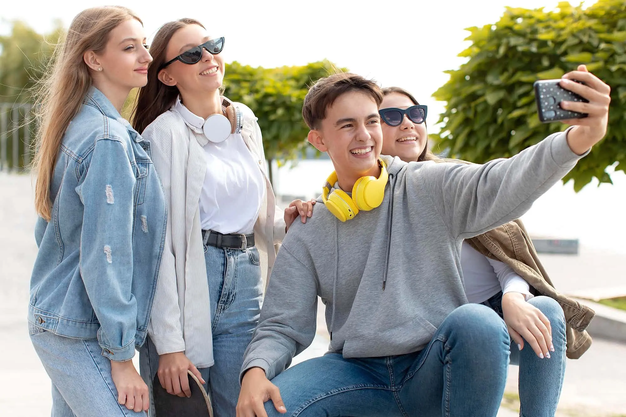 A group of four young people, outdoors in St. Augustine, FL, chat about different types of braces while taking a selfie—one holds the phone, two wear headphones around their necks, and trees with a walkway are visible behind them. The scene is linked to Happy Smiles Orthodontics.