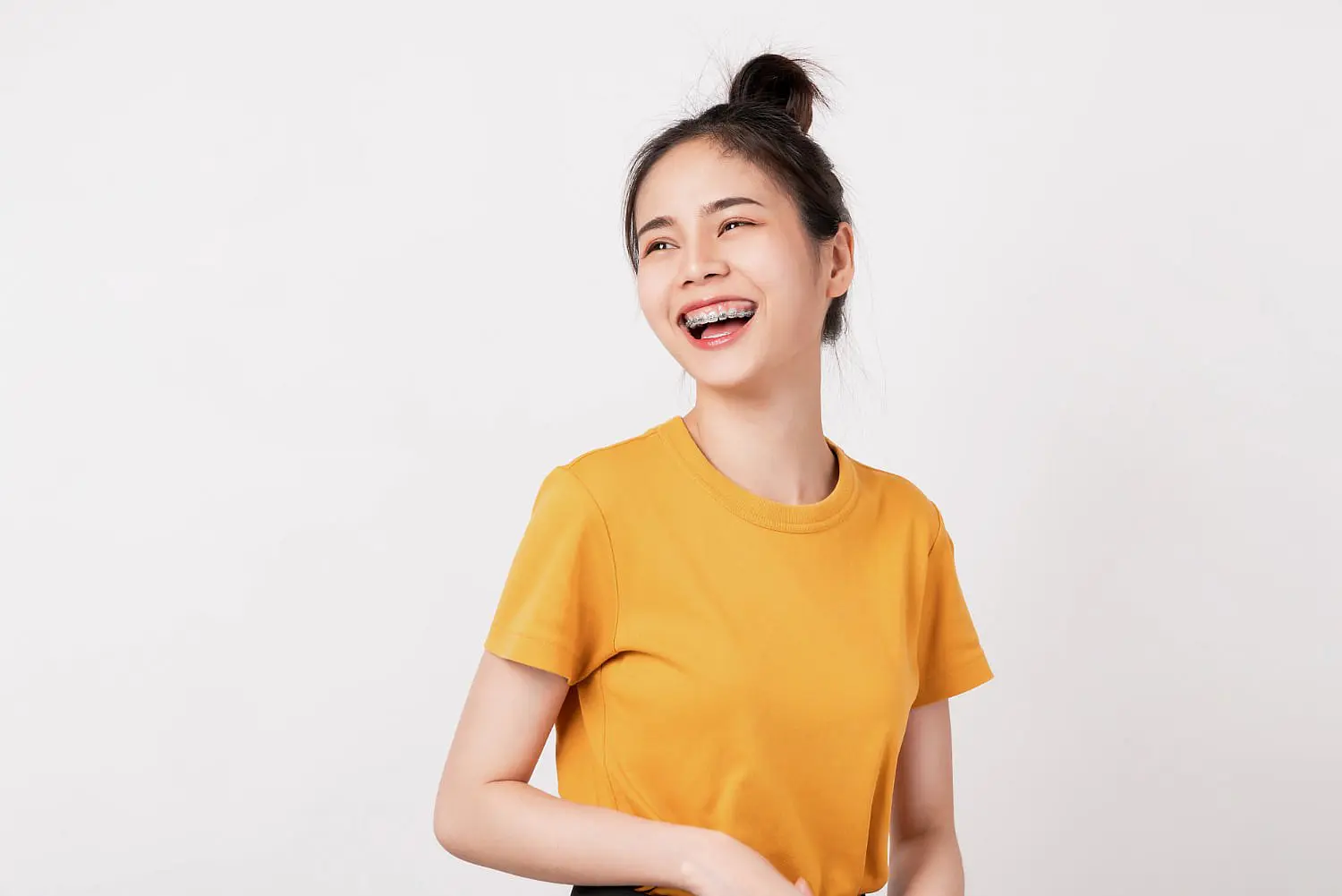 A young woman with long dark hair in a bun, wearing a yellow t-shirt and adult braces, smiles and gazes to the side against a plain white background at Happy Smiles Orthodontics in St. Augustine, FL.