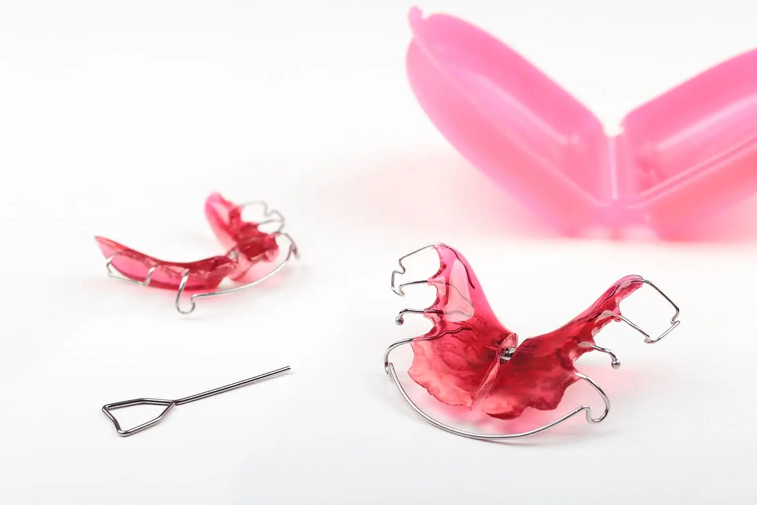 Displayed on a white surface are two pink orthodontic retainers, a metal adjustment tool, and an open pink plastic case—essential items recommended by Children’s Orthodontists at Happy Smiles Orthodontics in St. Augustine, FL for use with functional appliances.