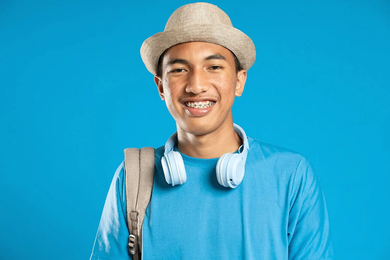 Smiling in front of a blue background, a young person with braces from Happy Smiles Orthodontics in St. Augustine, FL wears a hat, blue shirt, headphones around their neck, and carries a backpack.