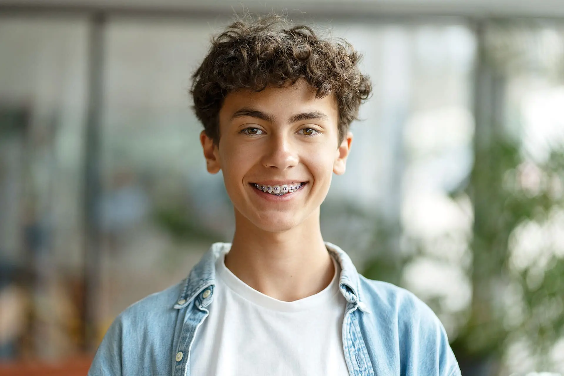 A teen with curly hair, wearing a white t-shirt and denim shirt, smiles confidently with braces against a blurred indoor background at Happy Smiles Orthodontics in St. Augustine, FL—an ideal image for those interested in Invisalign vs Braces.