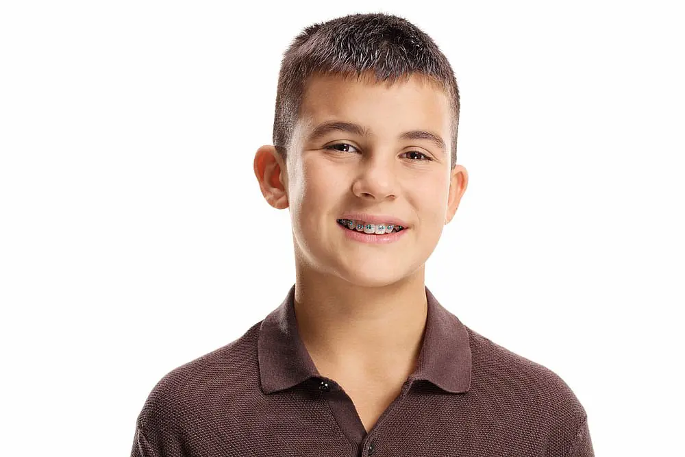 A smiling boy with short dark hair and traditional braces, dressed in a brown polo shirt and standing against a plain white background, showcases the results achieved at Happy Smiles Orthodontics in St. Augustine, FL — a trusted children’s orthodontist committed to creating confident smiles.