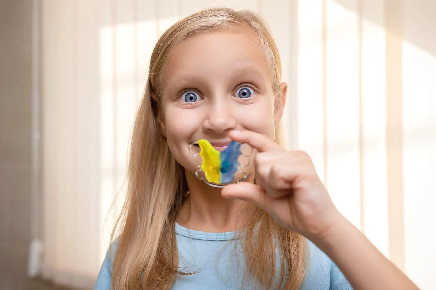 A young girl with long blonde hair proudly smiles indoors, showing off a colorful yellow-and-blue striped retainer—thanks to the expert care at Happy Smiles Orthodontics in St. Augustine, FL.