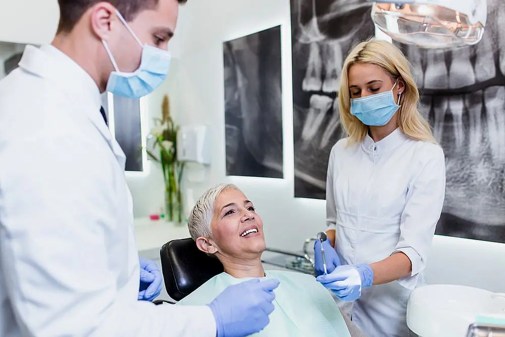 At Happy Smiles Orthodontics in St. Augustine, FL, a woman smiles in a modern dental chair as two masked and gloved dental professionals prepare tools for her adult orthodontics treatment.