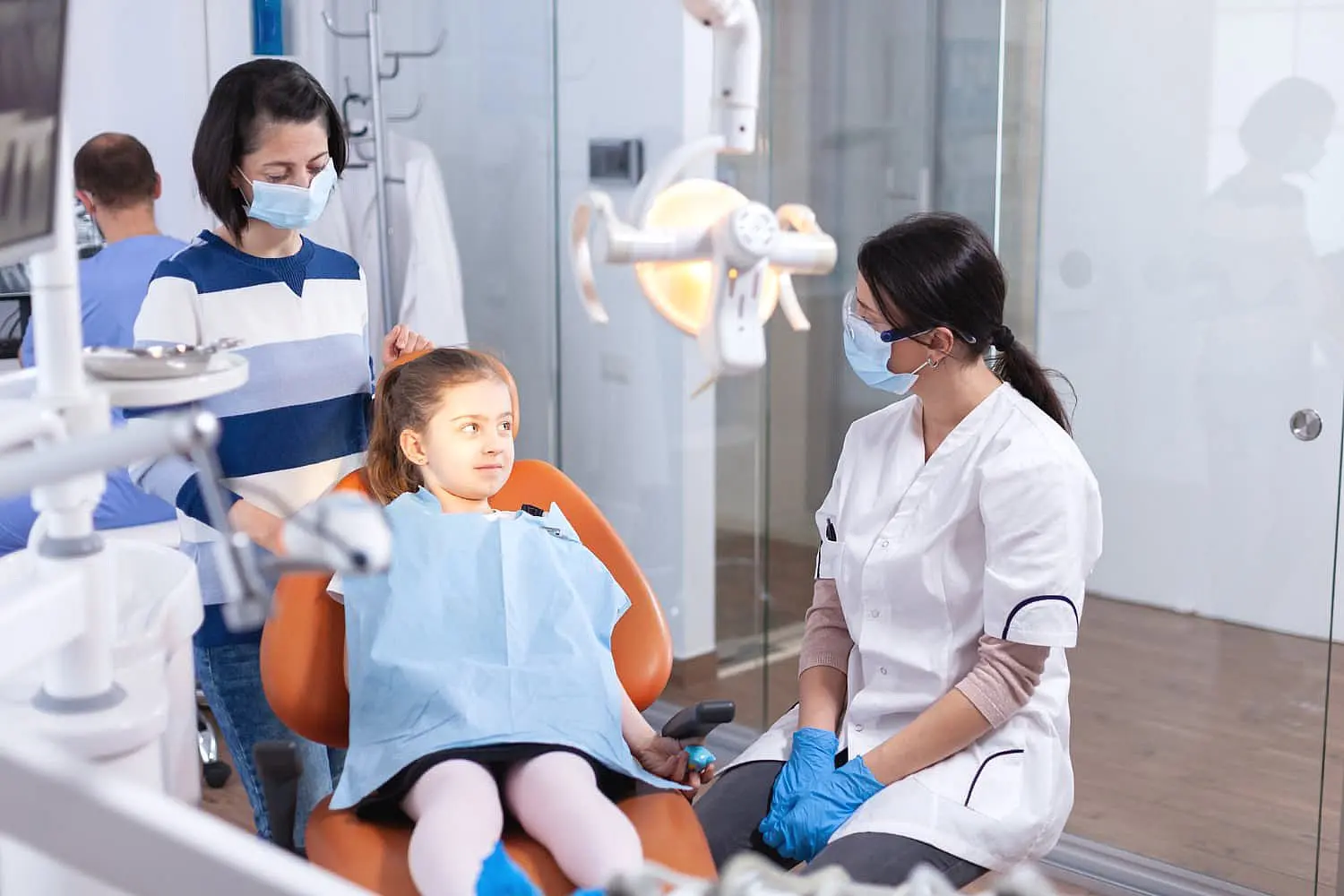 At Happy Smiles Orthodontics in St. Augustine, FL, a children’s orthodontist wearing a mask and gloves talks to a young girl sitting in the dental chair with her guardian in a modern office.