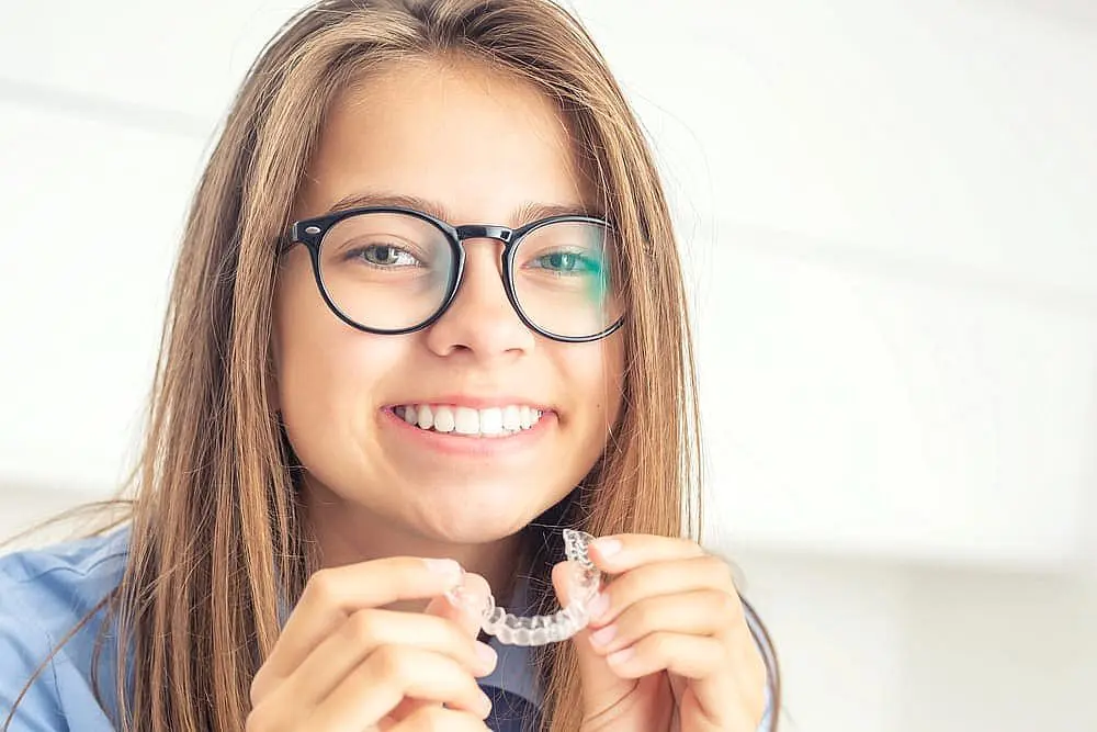 At Happy Smiles Orthodontics in St. Augustine, FL, a young person with long hair and glasses confidently smiles while holding clear aligners in both hands after visiting the children’s orthodontist.
