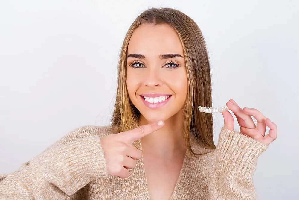 A woman smiles while holding a clear dental aligner and pointing to her teeth, showcasing how an Invisalign Payment Plan at Happy Smiles Orthodontics in St. Augustine, FL makes achieving a confident, affordable smile possible.
