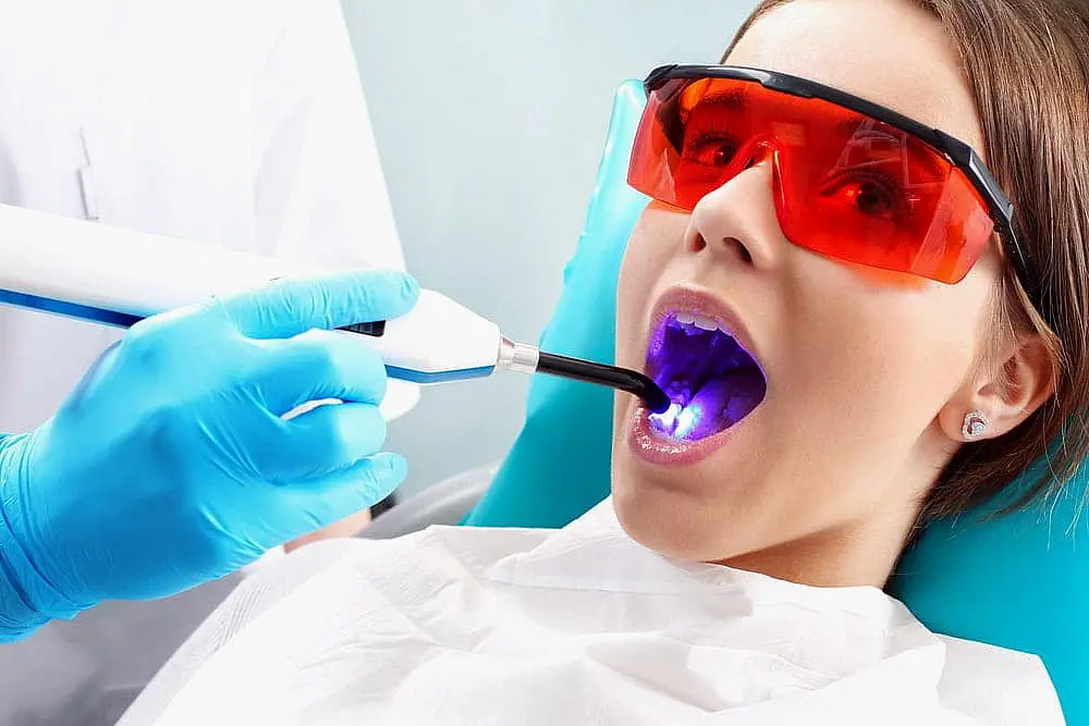 At Happy Smiles Orthodontics in St. Augustine, FL, a clinician in blue gloves uses a curing light during dental procedures—such as Invisalign vs braces adjustments—while the patient wears protective glasses.
