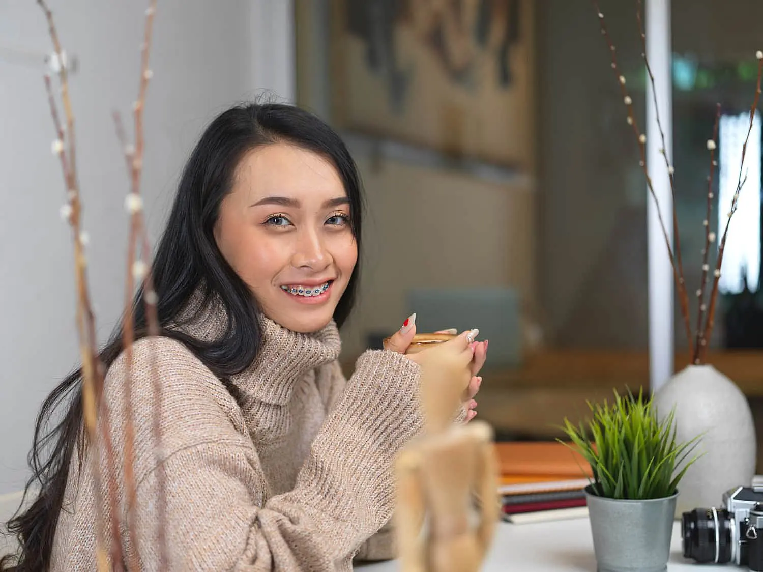 At Happy Smiles Orthodontics in St. Augustine, FL, a woman with long dark hair and adult braces sits at a table wearing a beige sweater, holding a mug beside plants and a camera.