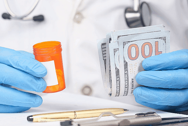 At Happy Smiles Orthodontics in St. Augustine, FL, an individual wearing blue gloves holds a pill bottle and a stack of hundred-dollar bills, highlighting the connection between healthcare costs and flexible options like an Invisalign Payment Plan.